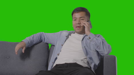 Studio-Shot-Of-Young-Man-Sitting-On-Sofa-Talking-On-Mobile-Phone-Against-Green-Screen-