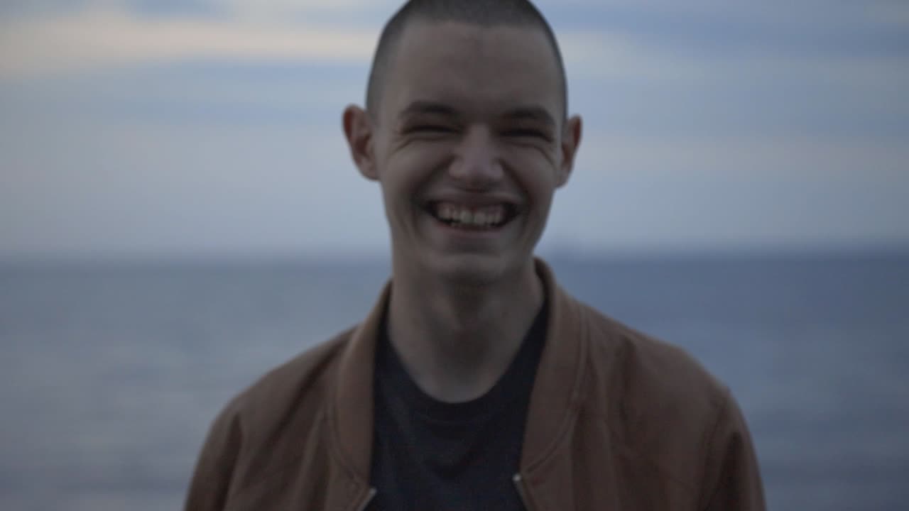Young Man Laughing While Looking Directly At The Camera Isolated On Blurry Blue Sea And Sky Background - Closeup Shot