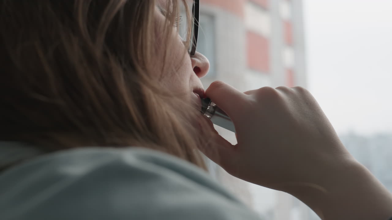 Caucasian woman at window contemplative closeup, glasses catching soft morning light, gentle smile and distant gaze, fingers near lips suggesting thoughtful pause, intimate indoor portrait with urban