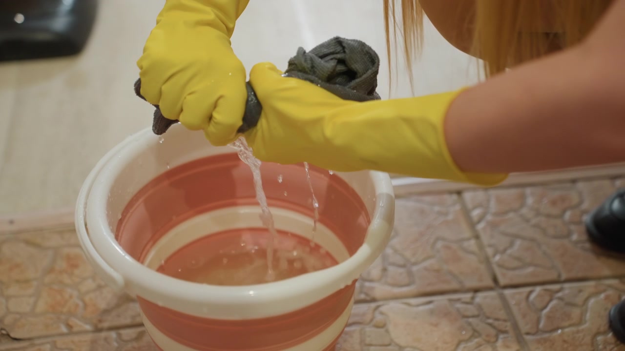Woman squeezing mop into bucket, yellow rubber gloves wringing gray rag over pink bucket on tiled floor, soapy water dripping, closeup hands pressing rag, domestic cleaning chore in home, calm