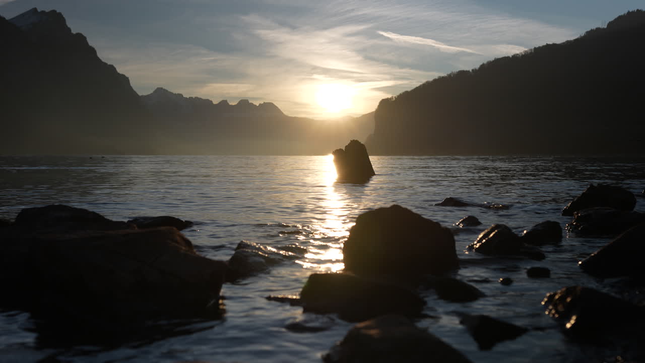 Sunset over Walensee, Switzerland, casting golden light over the rippling lake and surrounding mountains. Silhouetted rocks in the foreground enhance the serene and dramatic alpine scenery.