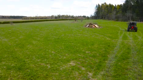 flying over grassy countryside field while loaded farm vehicle hauls dry branches, wood into large pile in the middle of a pastureland. homestead spring work. forest edge and farmland in background