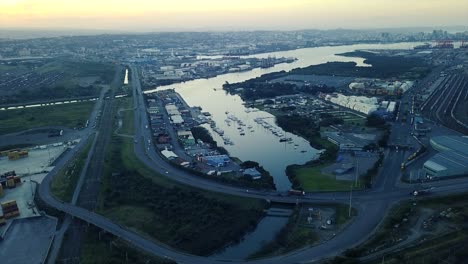 Aufnahmen-Von-Sonnenuntergang-Mit-Blick-Auf-Yachten,-Die-Mit-Verkehr-Auf-Einer-Straße-Festgemacht-Sind,-Und-Schiffscontainern,-Die-Von-Lastwagen-Zu-Schiffen-Transportiert-Werden