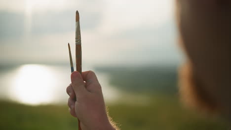 a close-up of a hand holding two paintbrushes one small and one very small against a sunny, blurred background. the soft lighting emphasizes the tools of the artist, capturing a moment of creative