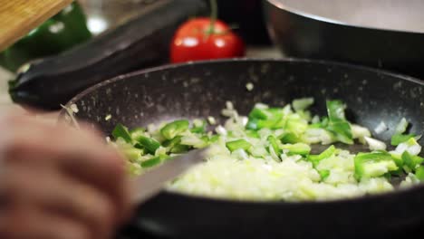 Scraping-chopped-white-onions-with-a-knife-from-a-wooden-chopping-board-into-a-black-frying-pan-with-chopped-green-sweet-peppers