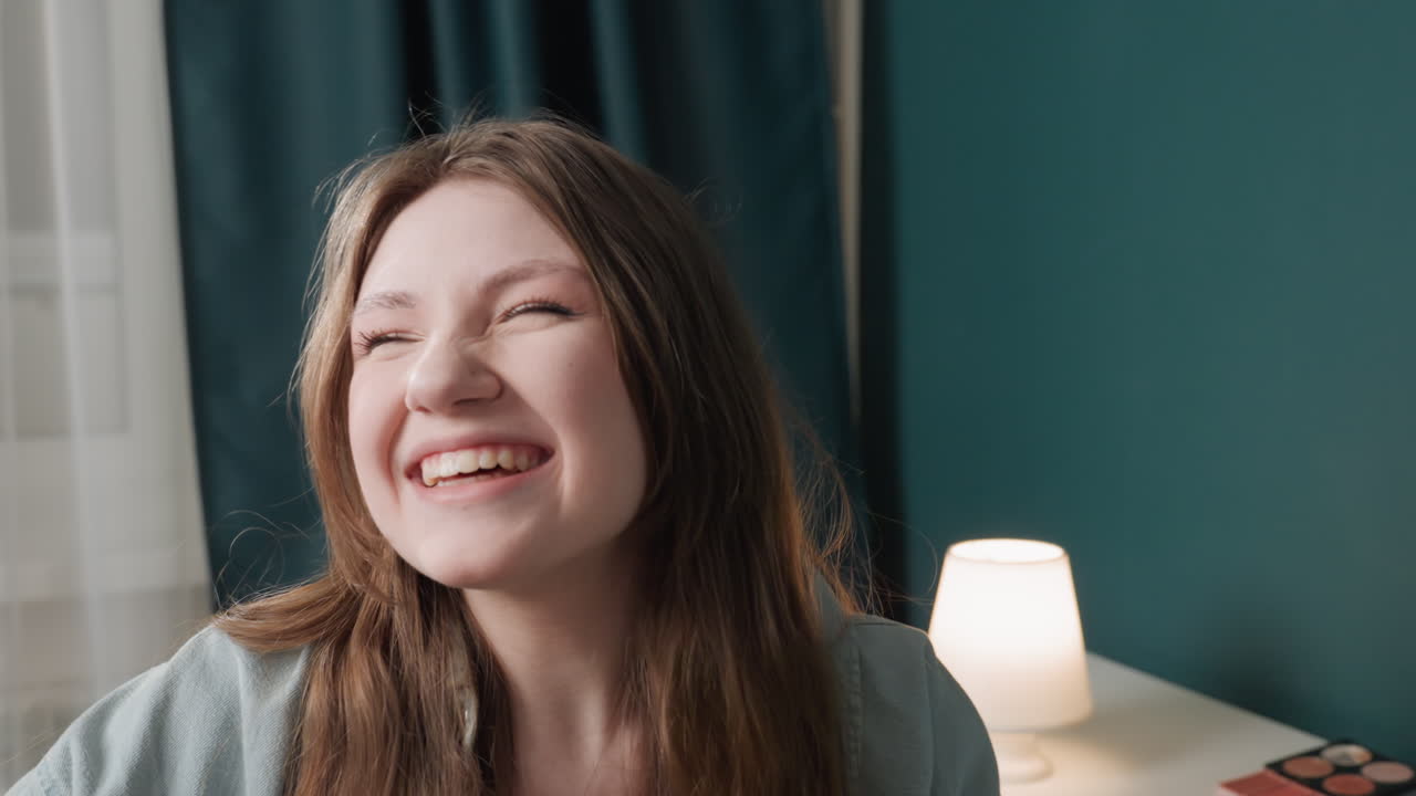 Giovane donna caucasica che ride candidamente in una scena accanto al letto, stanza luminosa e accogliente con lampada e palette di trucchi sulla scrivania, risata espressiva con mano sul mento e occhi chiusi, sorriso genuino ed energia positiva