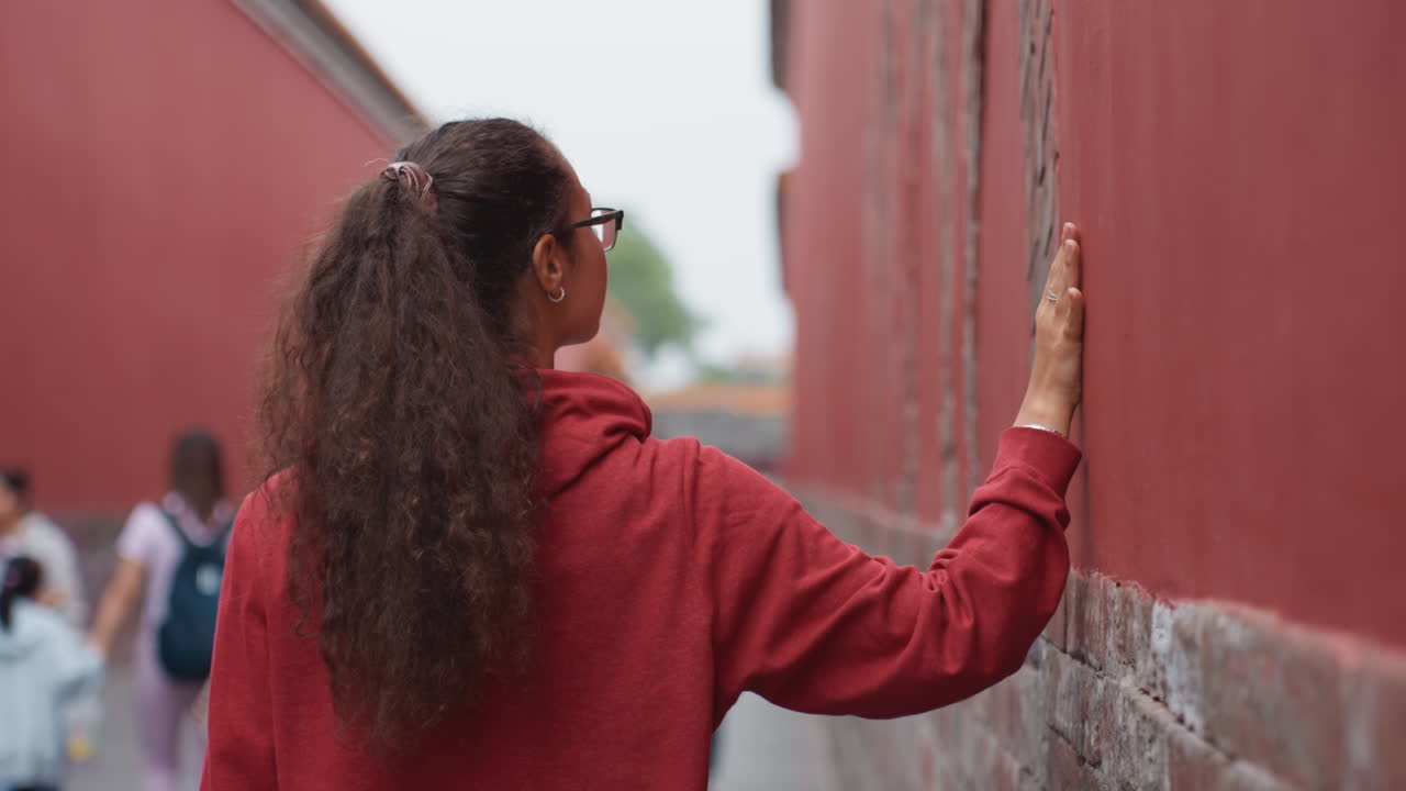 Donna che tocca un muro rosso in un vicolo, che indossa una felpa rossa con cappuccio e occhiali, capelli ricci legati a coda di cavallo, mano appoggiata su mattone consumato, pausa contemplativa di turista in mezzo a una folla distante, corridoio del tempio in prospettiva.