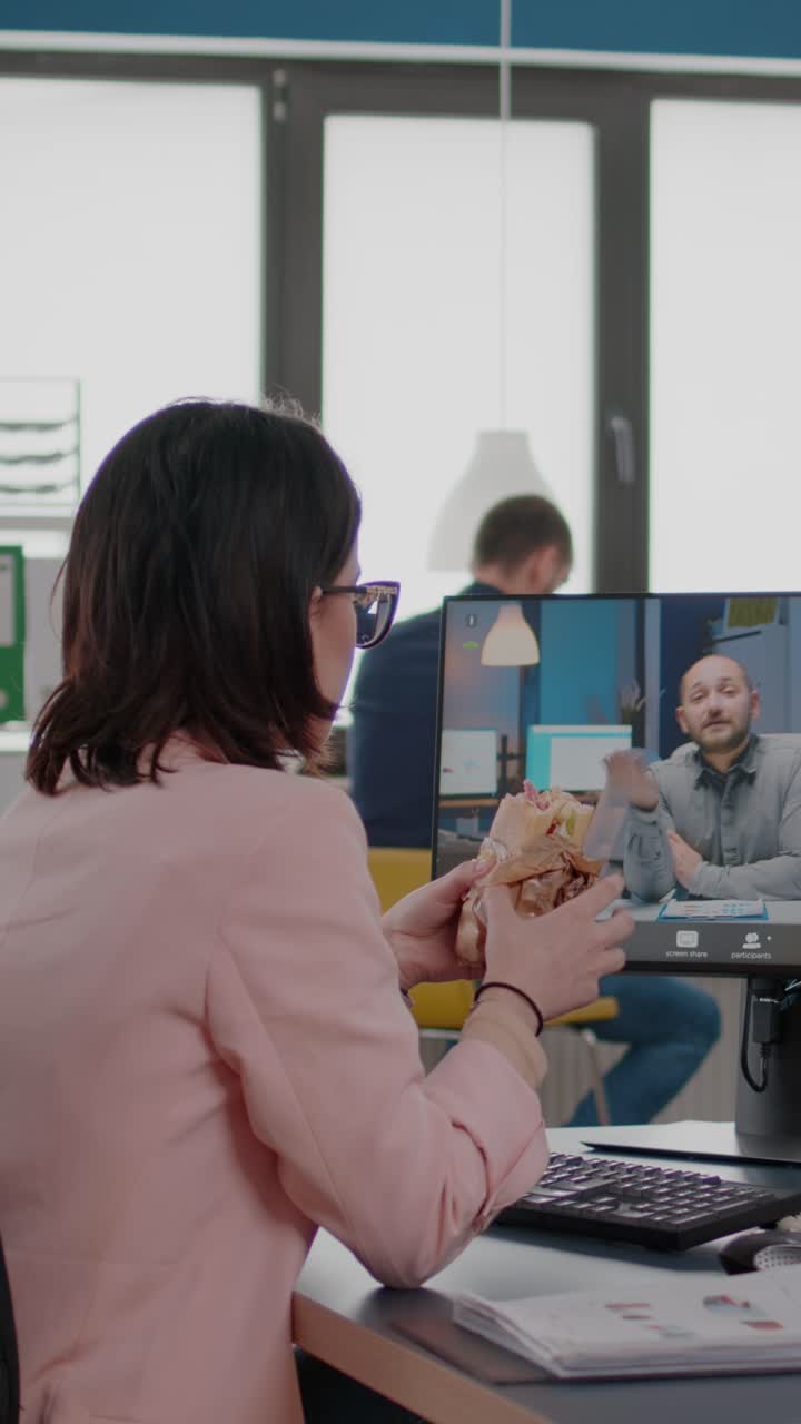 Vertical video: Entrepreneur woman sitting at desk in company office eating sandwich during online videocall conference