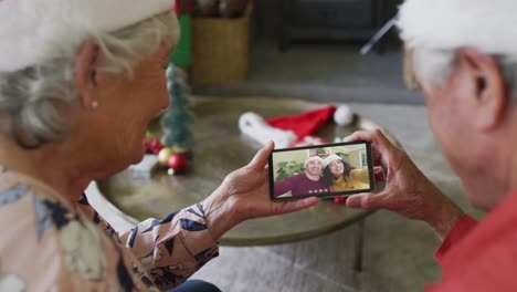 Pareja-Caucásica-Mayor-Usando-Un-Teléfono-Inteligente-Para-Una-Videollamada-Navideña-Con-Una-Pareja-Sonriente-En-La-Pantalla