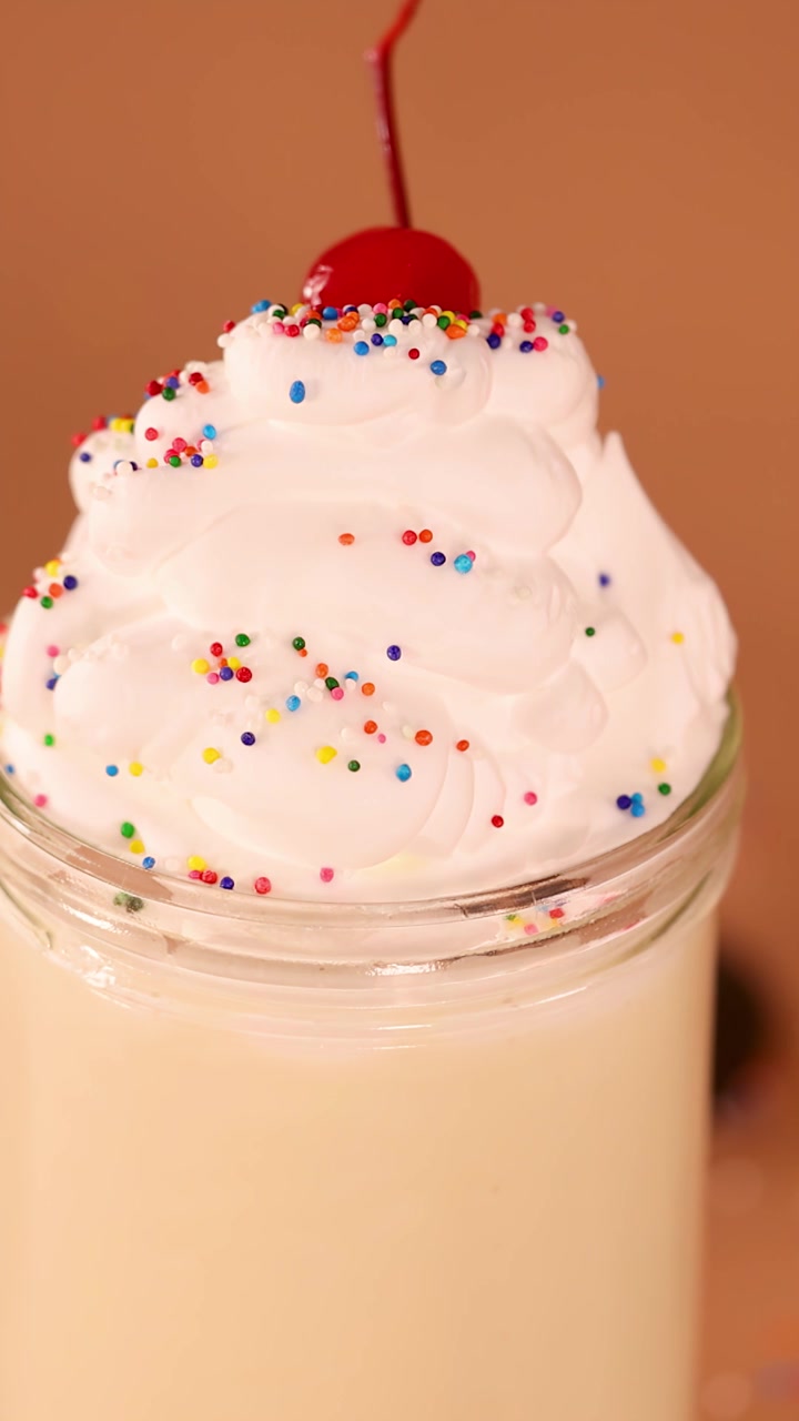 Primo piano al rallentatore di una ciliegia che scivola via da un milkshake alla vaniglia con panna montata su uno sfondo caldo.