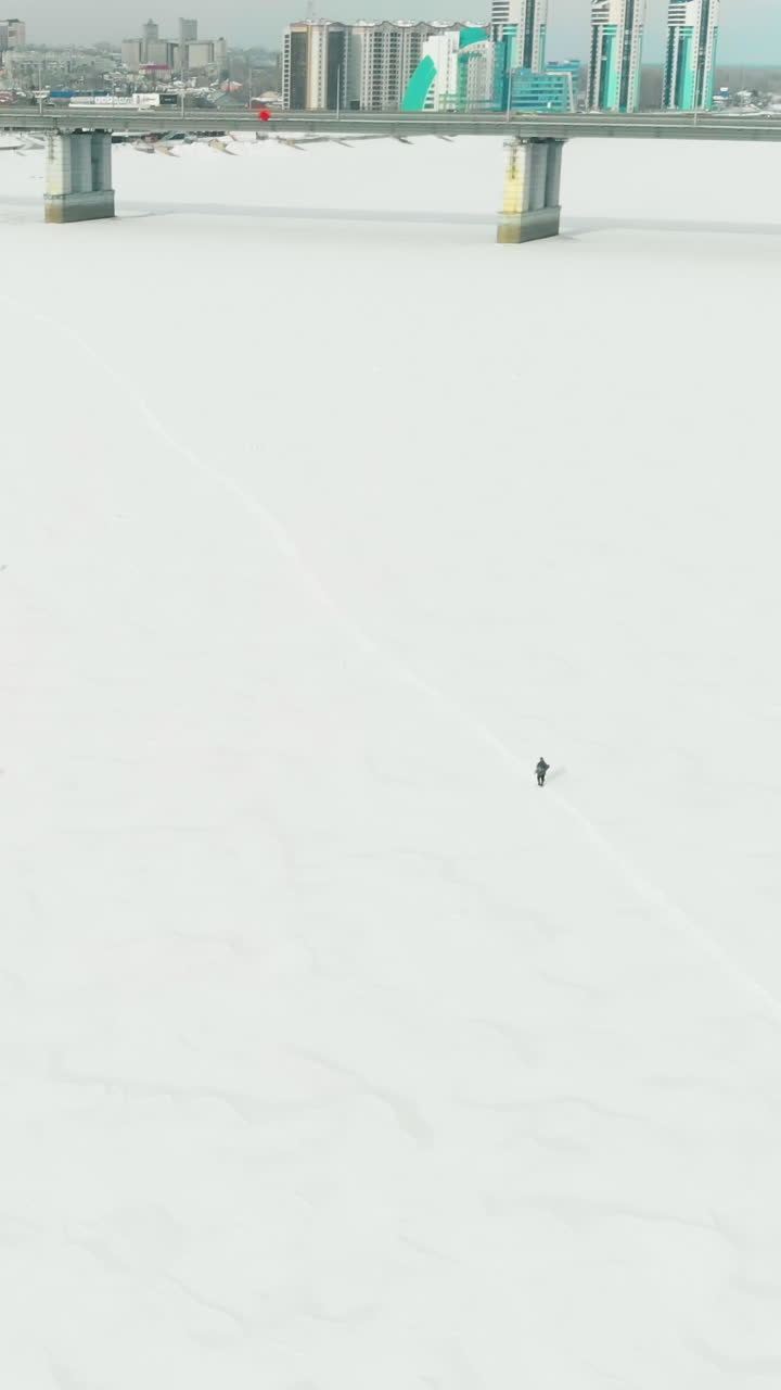 blurry person silhouette walks along snowy path on frozen river surface against grey bridge upper view