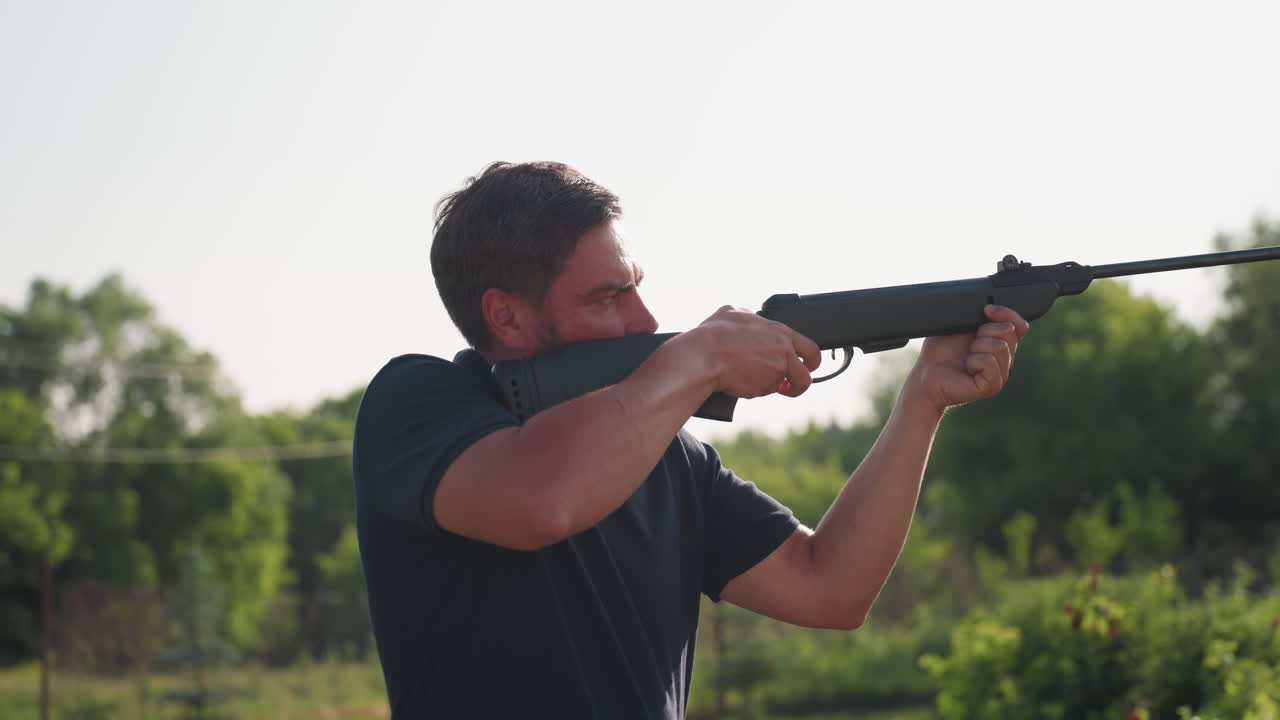 Gardener with pneumatic rifle aims and shoots to scare pests from crops in green field, focused stance in sunny rural garden with trees and fence, protective action during daylight