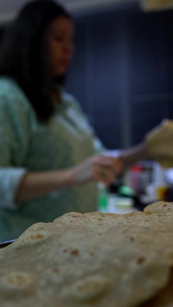 Woman in light green shirt preparing and cooking whole wheat chapati flatbread on stovetop. Close-up focus on golden-brown cooked bread with visible air pockets, traditional Indian cooking, warm kitch