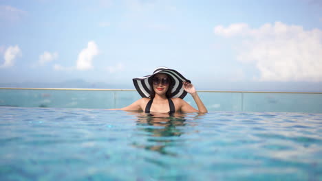 Beautiful-woman-with-a-pretty-smile-wearing-a-sun-hat-and-glasses-relaxes-in-the-clear-rippling-waters-of-a-lavish-infinity-pool-under-bright-blue-skies-with-white-clouds-and-the-sea-in-the-background