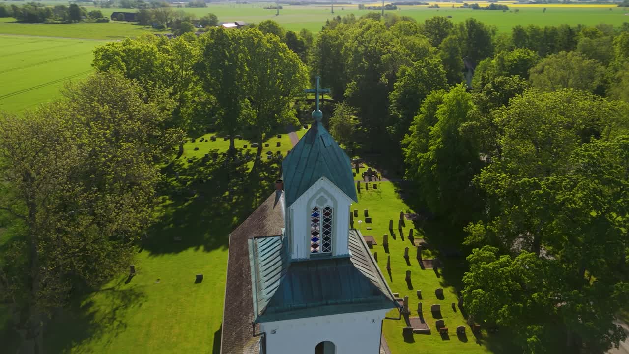 Parallaxen-Luftaufnahme, die den Kirchturm und Friedhof von Grinstad umkreist, umgeben von Wald und Ackerflächen im sonnigen ländlichen Schweden.
