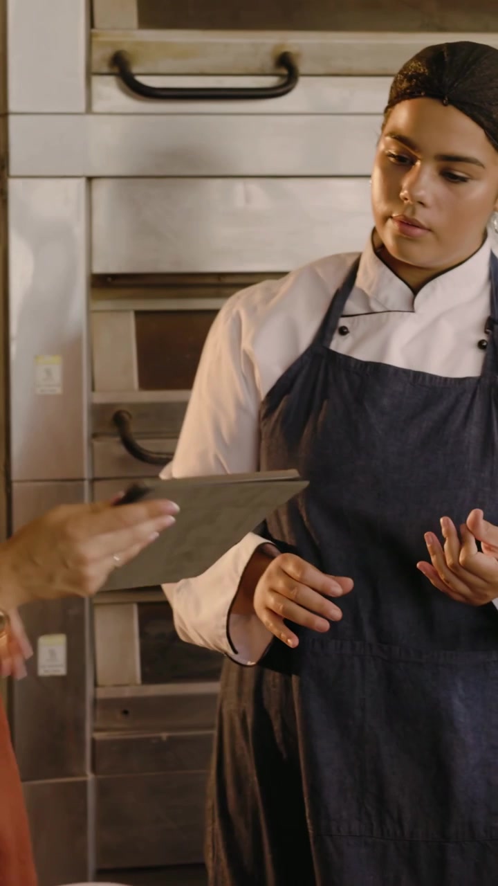 Video vertical: Chef llegando con chaqueta, recibiendo una tablet de un miembro del personal con camisa color óxido junto a los hornos, planificando