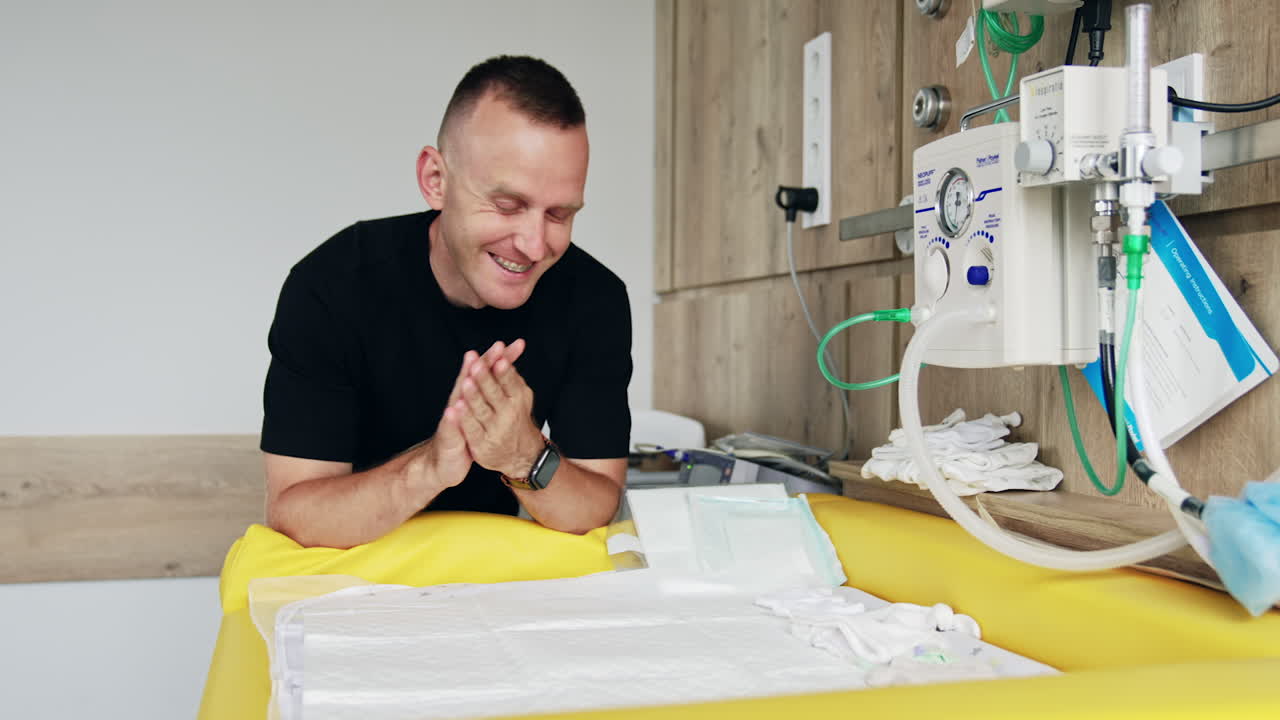 Happy expecting dad stands at the table with kid's clothes. Rejoicing man shows tiny white baby socks.
