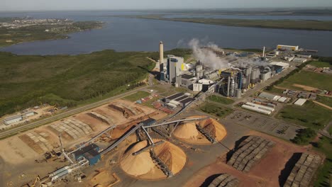 aerial view of paper mill factory with smoky chimneys front to river - sawdust piles and pipes transporting wood trunks