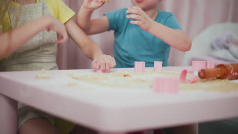 young siblings engaged in joyful baking session with girl pressing cookie cutter into dough while brother removes cut circles stacking shapes on pink table surface surrounded by flour together