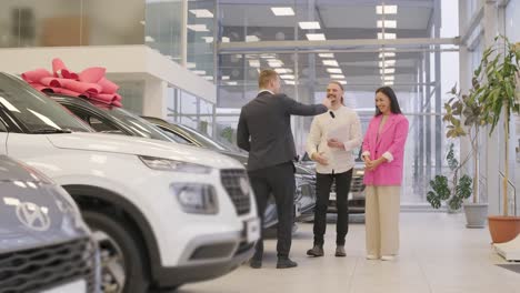 young car salesman showing to young couple new automobile at dealership salon.