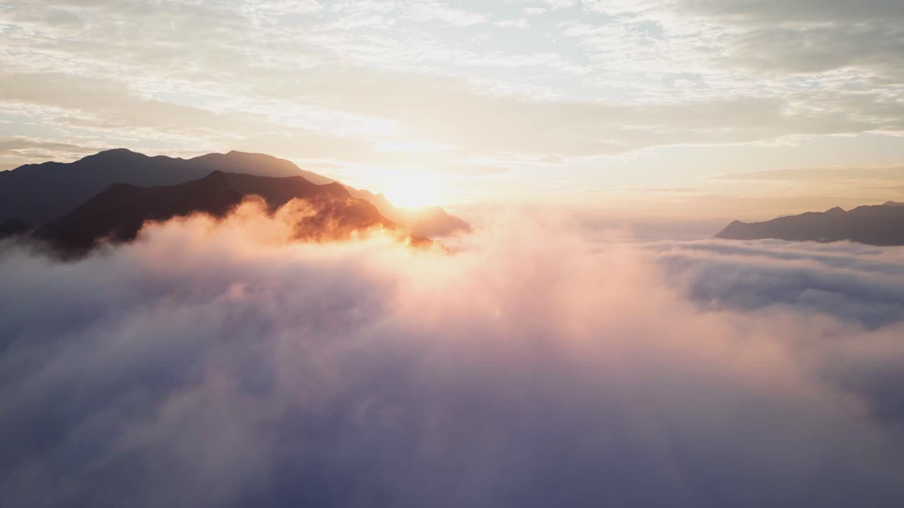 Riprese da drone catturano un'alba maestosa sopra uno spesso strato di nuvole che si estende su montagne aspre. Calda luce dorata si riflette su nuvole testurizzate mentre vette lontane emergono dalla nebbia.