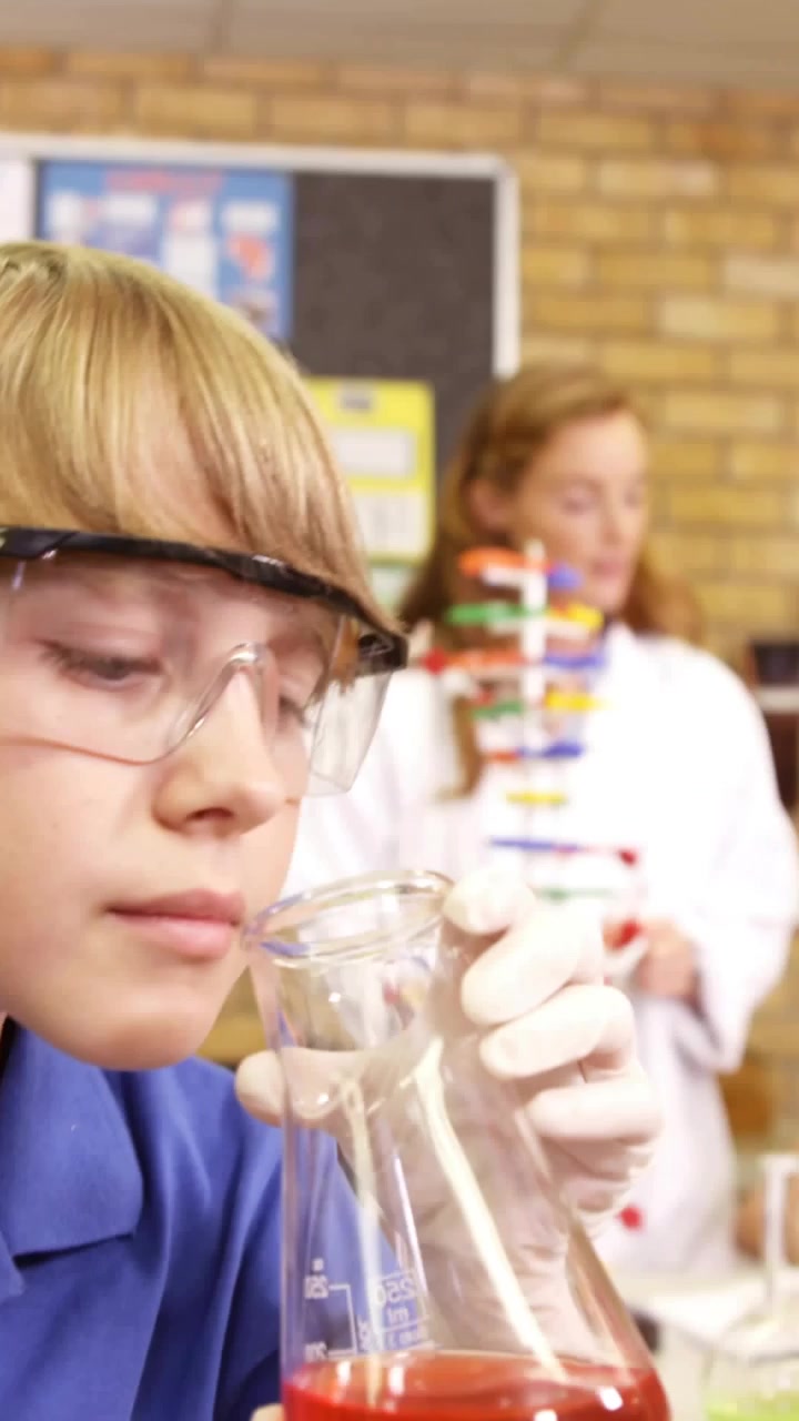 Vertical video: Gloved hand lifting flask causing code overlay as pupil checking red liquid in lab