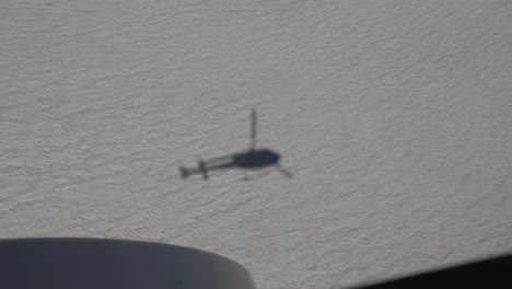 view from a helicopter down on its own shadow in the snow while flying into the mountains on a clear summer day in 4k