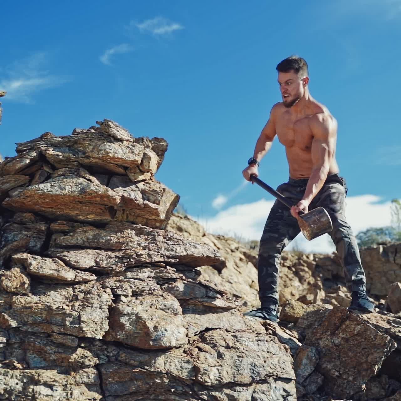 Athletic man exercises with a big hammer in canyon. Muscular man standing on rock and breaking stones with a sledgehammer under the blue sky. Slow motion.