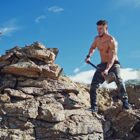 athletic man exercises with a big hammer in canyon. muscular man standing on rock and breaking stones with a sledgehammer under the blue sky. slow motion.