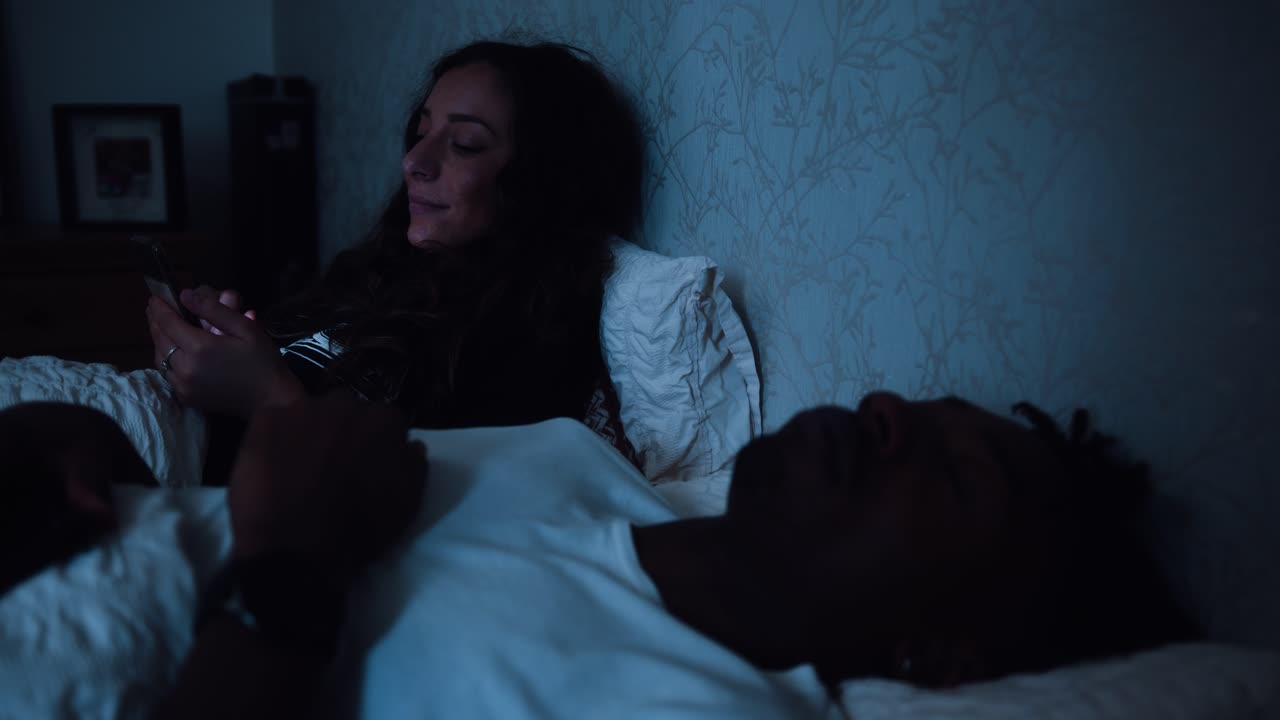 mujer se desplaza en su teléfono mientras su pareja duerme en la cama junto a ella tarde en la noche