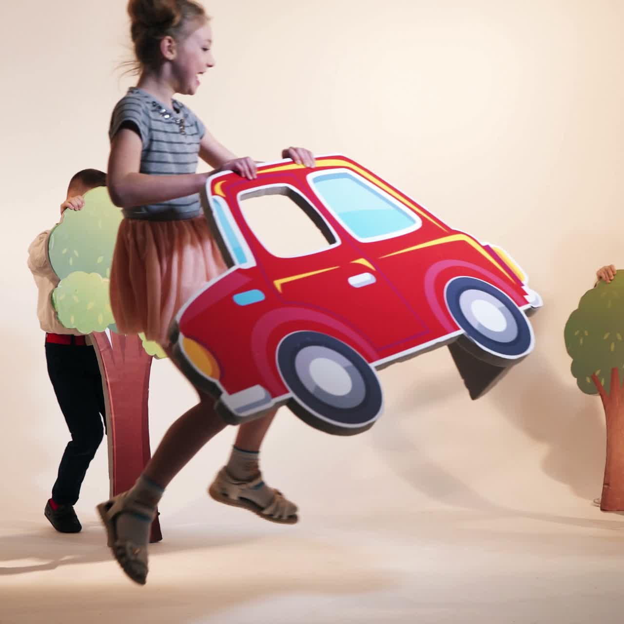 funny children are running with different decorations in the form of cars, trees and bushes on a studio background