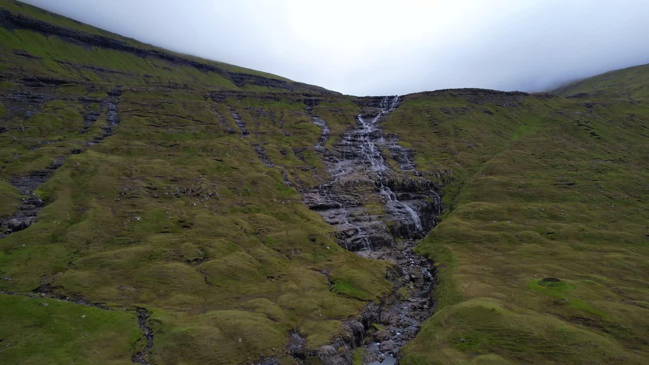 toma aérea en un río que fluye sobre un paisaje volcánico verde y brumoso en la isla de streymoy, islas feroe