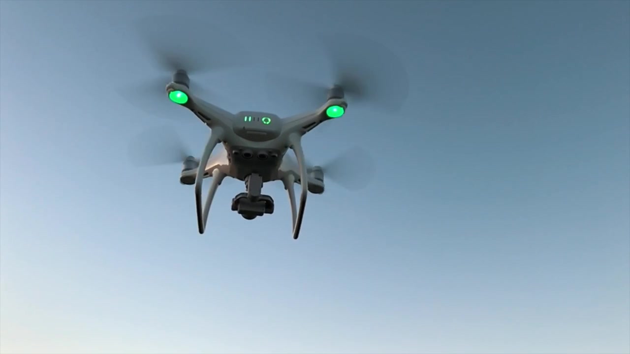 Drone quadricottero bianco che plana e ruota sul posto con sfondo di cielo blu e luci lampeggianti.