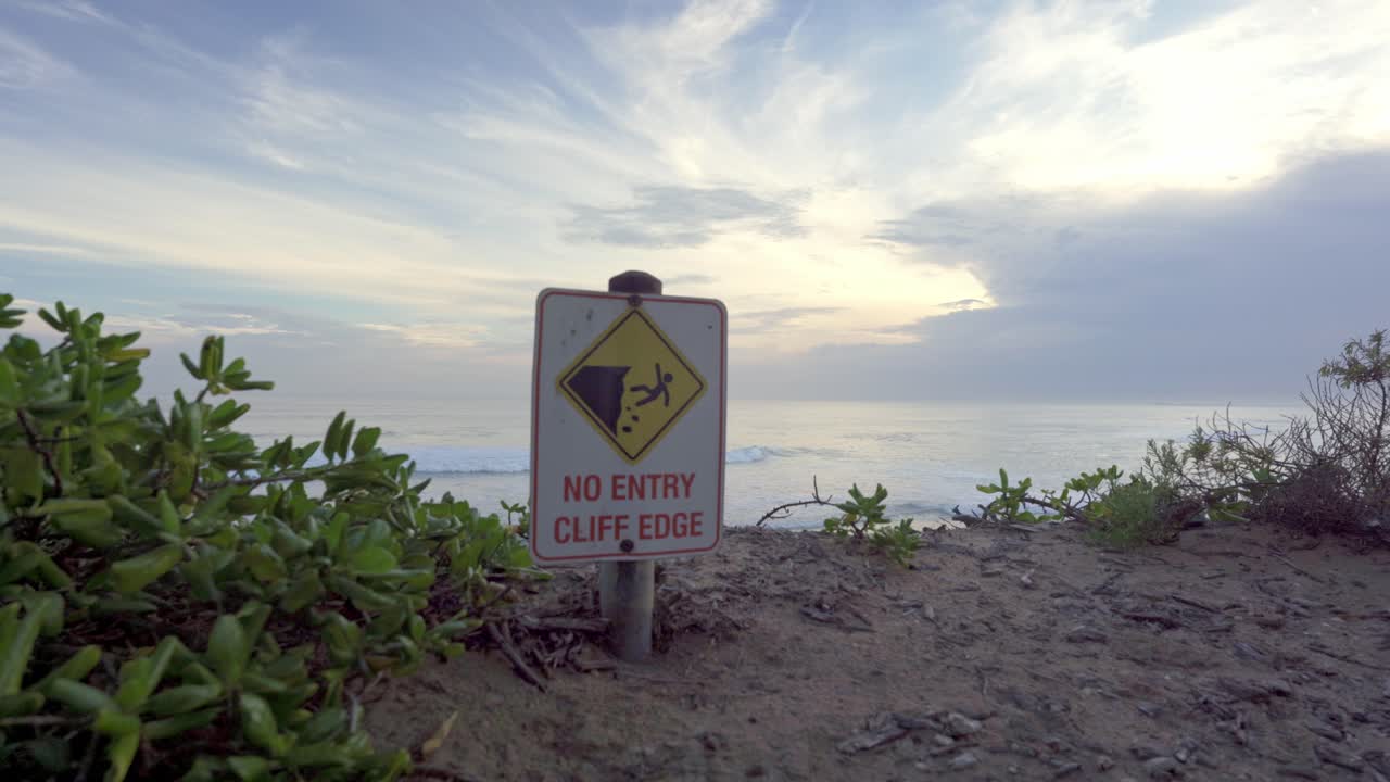 Segnale di avviso con la scritta "Divieto di accesso - Bordo della scogliera" su una falesia costiera sabbiosa sopra l'oceano a Bellarine Peninsula vicino a Melbourne, Victoria, Australia, con onde e nuvole al tramonto