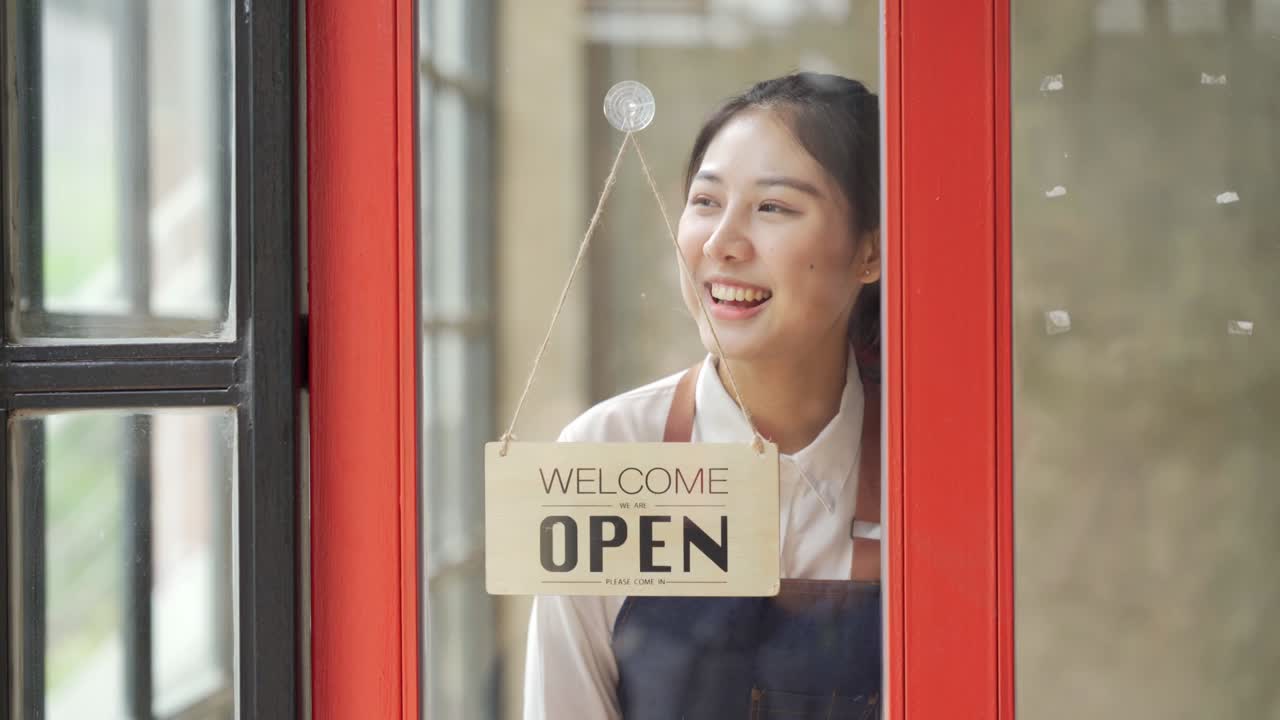 Young asian woman hanging an open sign on the window of a cafe, female owner of coffee shop or restaurant walking up to door and turning round sign to open.