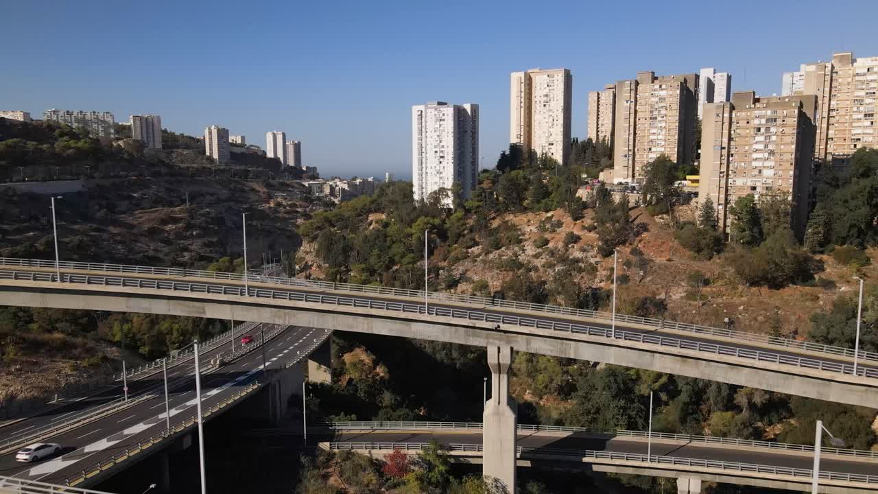 Volando dritto attraverso un complesso sistema di strade, incroci e curve sulle colline di Haifa, con alti edifici circondati da alberi e un orizzonte con cielo sereno in una giornata di sole, Israele.