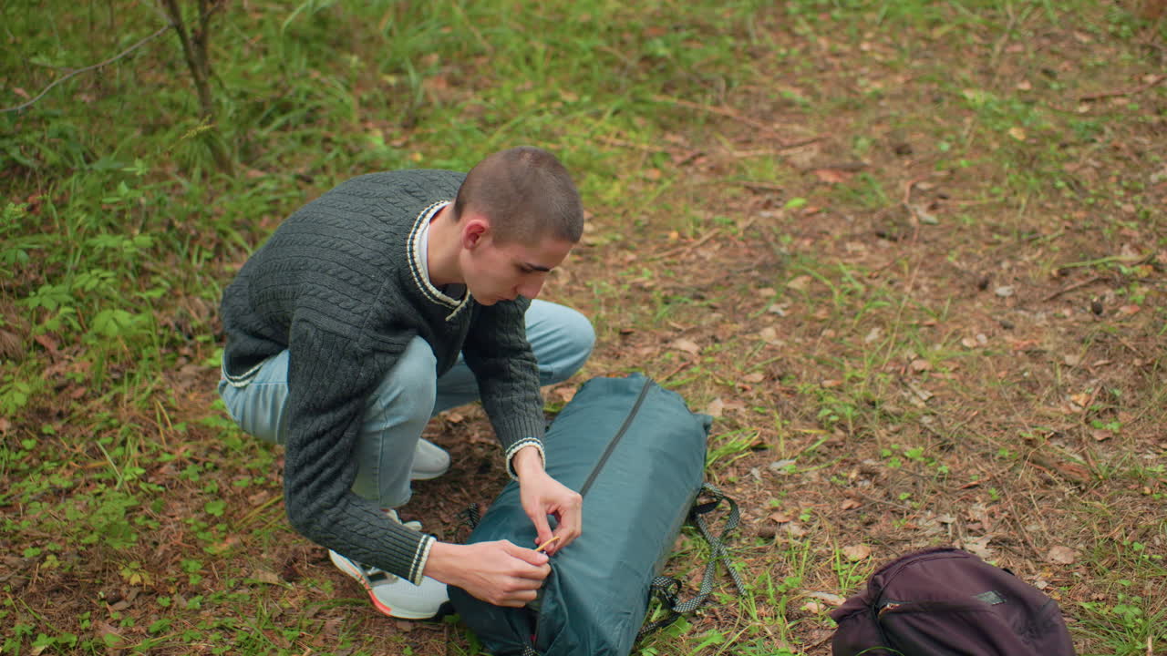 overhead view of young man in green knitted sweater crouched in woodland opening tent bag while dark backpack lies nearby on forest ground surrounded by dry leaves and green undergrowth