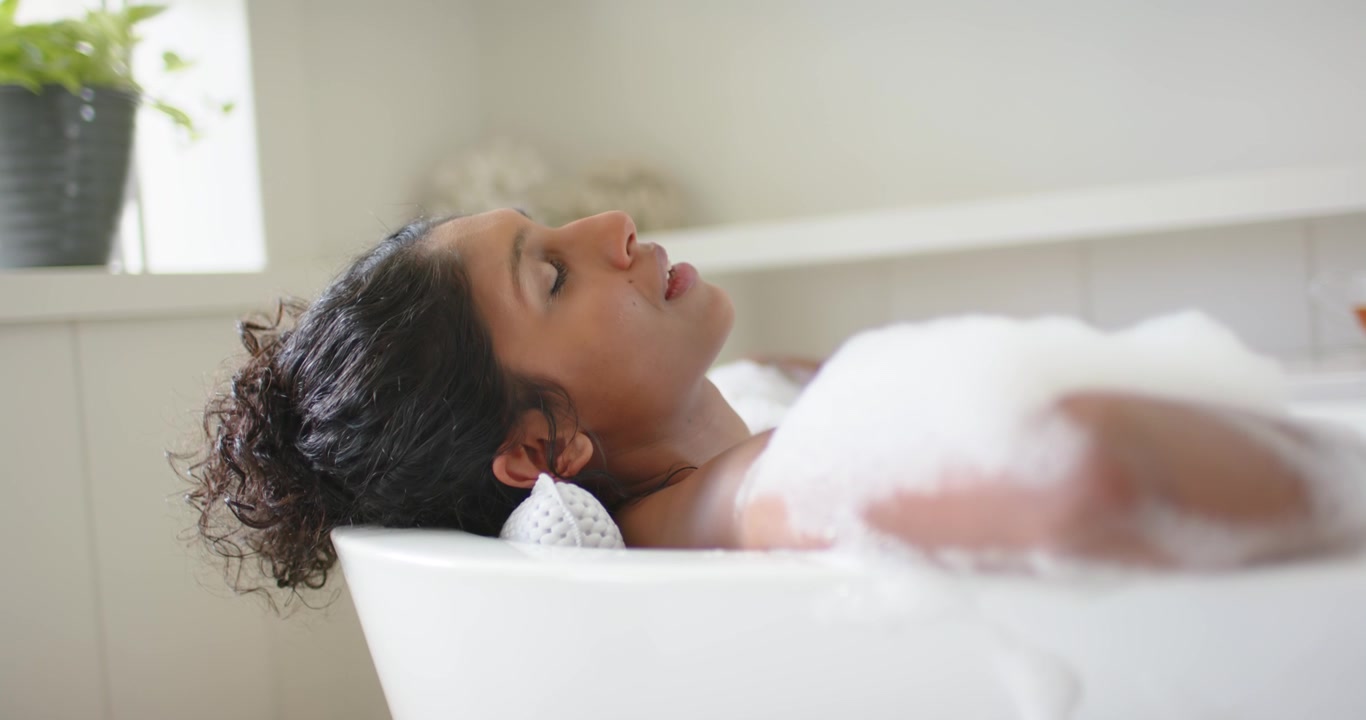 Settling into foam African American woman reclining in home tub moving arm to rest on pillow