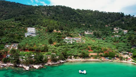 Casa-Junto-Al-Mar-Cerca-De-La-Playa-Dorada-Con-Barcos-Flotando-En-Aguas-Cristalinas-Turquesas,-Vegetación-Verde-Exuberante,-Plantación-De-Olivos,-Isla-De-Thassos,-Grecia