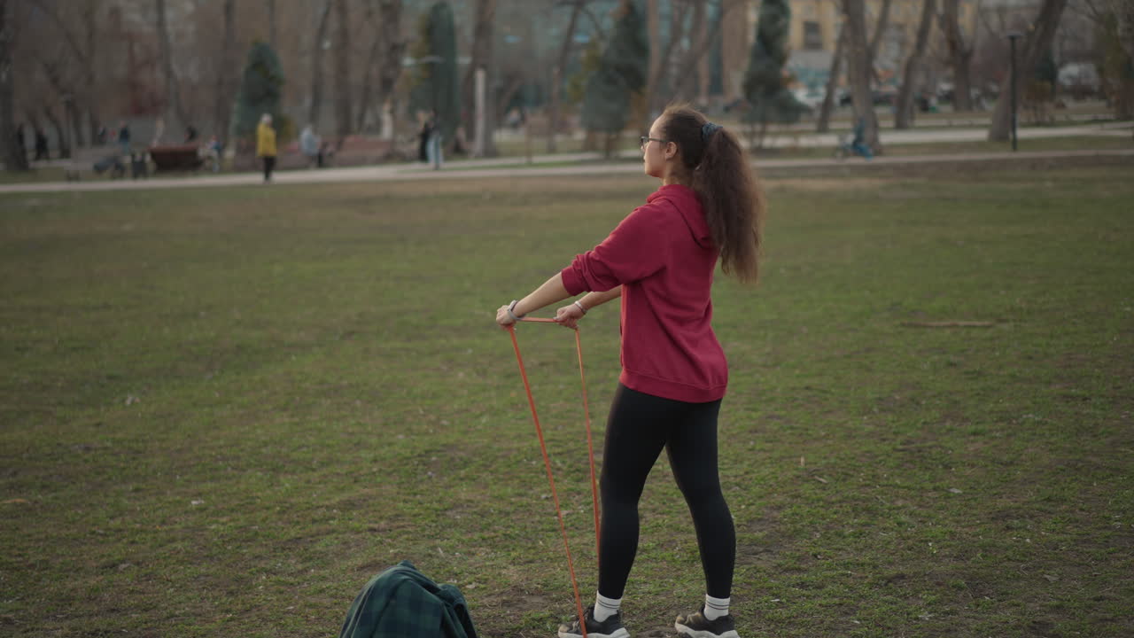 White Woman Training With Resistance Band In Public Park Among Walkers, Dynamic Posture And Full Body Pull, Red Hoodie And Ponytail, Backpack On Ground, Brisk Tempo And Social Urban Backdrop