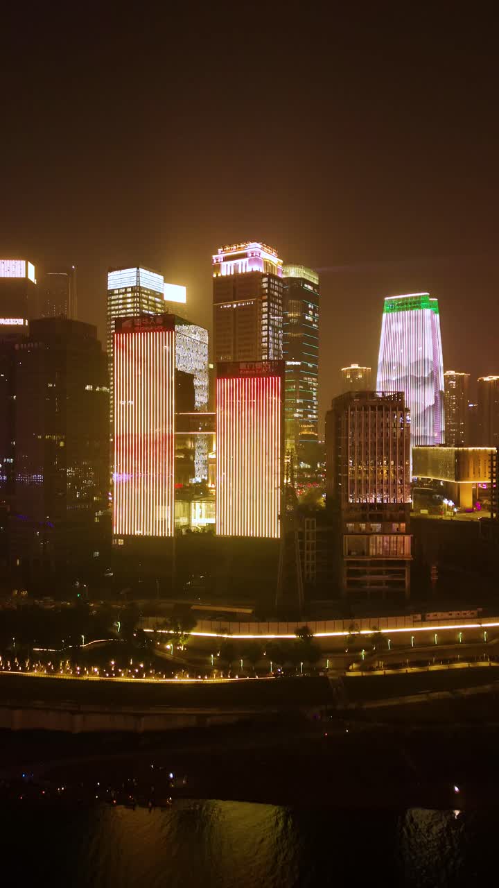 Vertical aerial shot of Chongqing's illuminated skyline, featuring modern skyscrapers with colorful LED displays reflecting on the river below. China