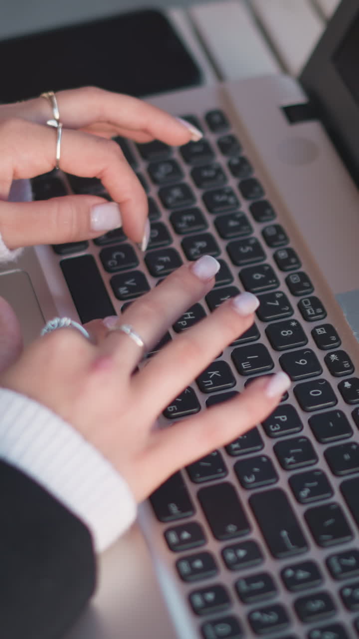 primer plano de manos adornadas con delicados anillos y uñas pulidas escribiendo en el teclado de la computadora portátil, reflejadas en la pantalla, las cómodas mangas del suéter añaden calor, con el teléfono inteligente cerca en la mesa de madera