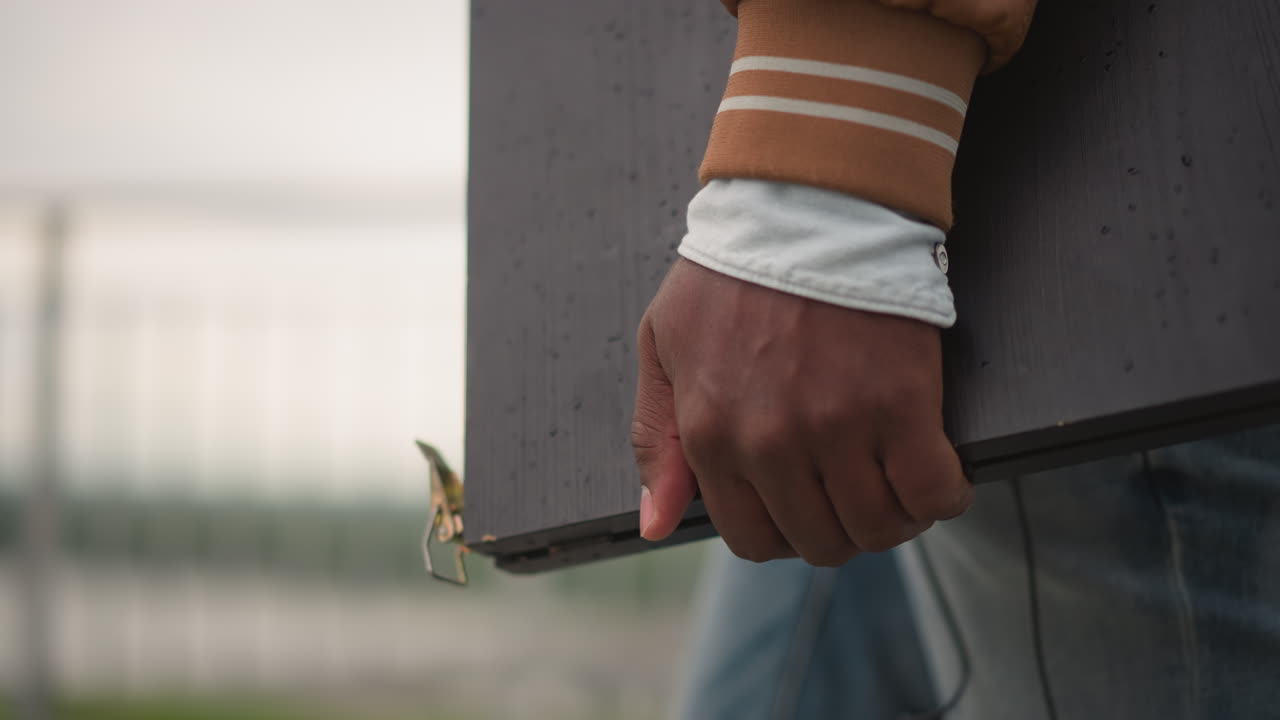 closeup hand holding dj case with raindrops, brown cuff and denim blurred waterfront railing in background, detailed metal clasp and worn texture, solitary musician preparing equipment, moody