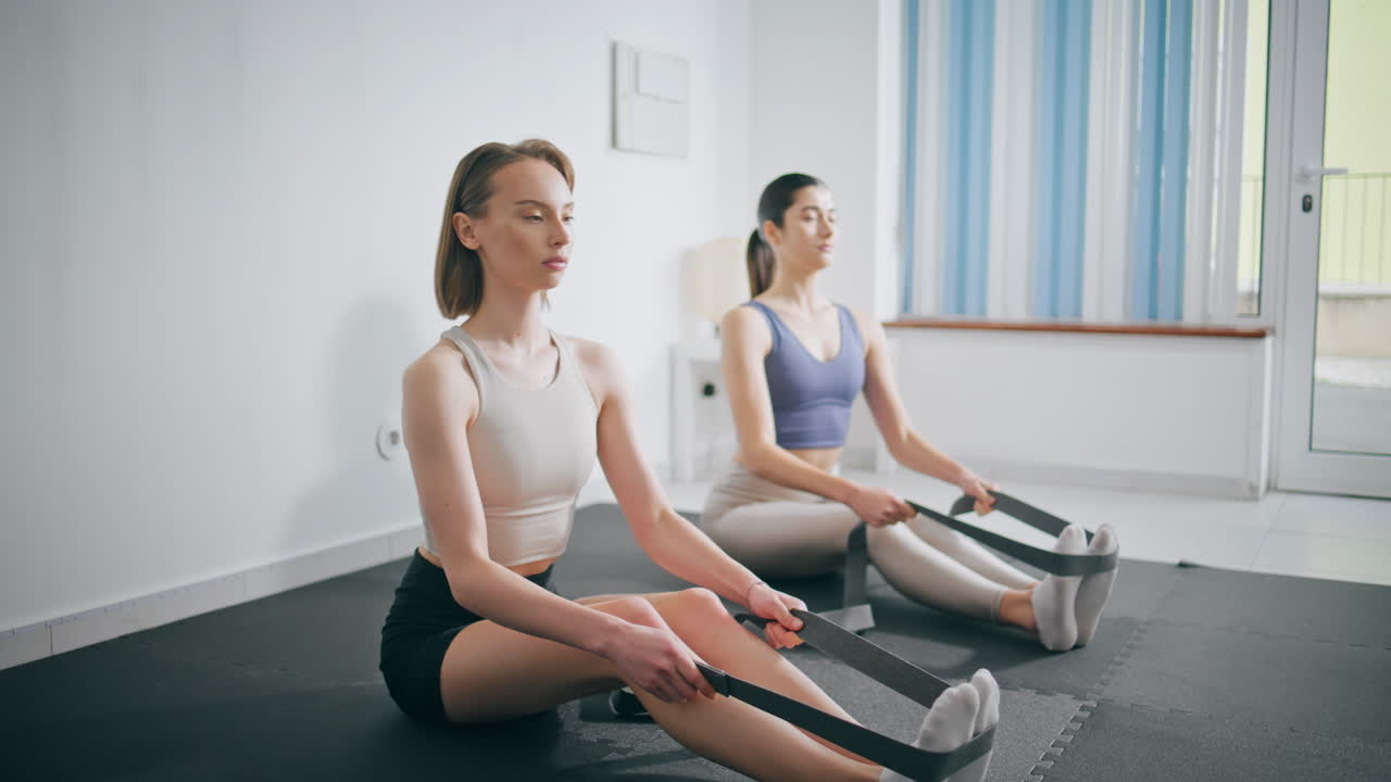 Athletic girls stretching body on yoga mat. Two women using fitness elastic band