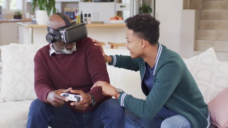 happy african american father using vr headset and gamepad with adult son at home, slow motion