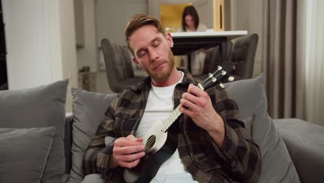 Confident-and-calm-blond-man-with-stubble-in-a-checkered-brown-shirt-plays-on-a-black-musical-string-instrument-ukulele-while-sitting-on-a-gray-sofa-while-his-wife-works-at-the-table-in-a-modern-apartment-during-the-day