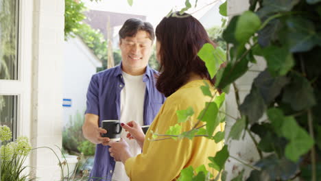 Mujer-Asiática-Madura-Plantando-Contenedores-De-Madera-En-El-Jardín-De-Verano-Mientras-El-Marido-Trae-Su-Bebida-Caliente-En-Una-Taza---Filmada-En-Cámara-Lenta