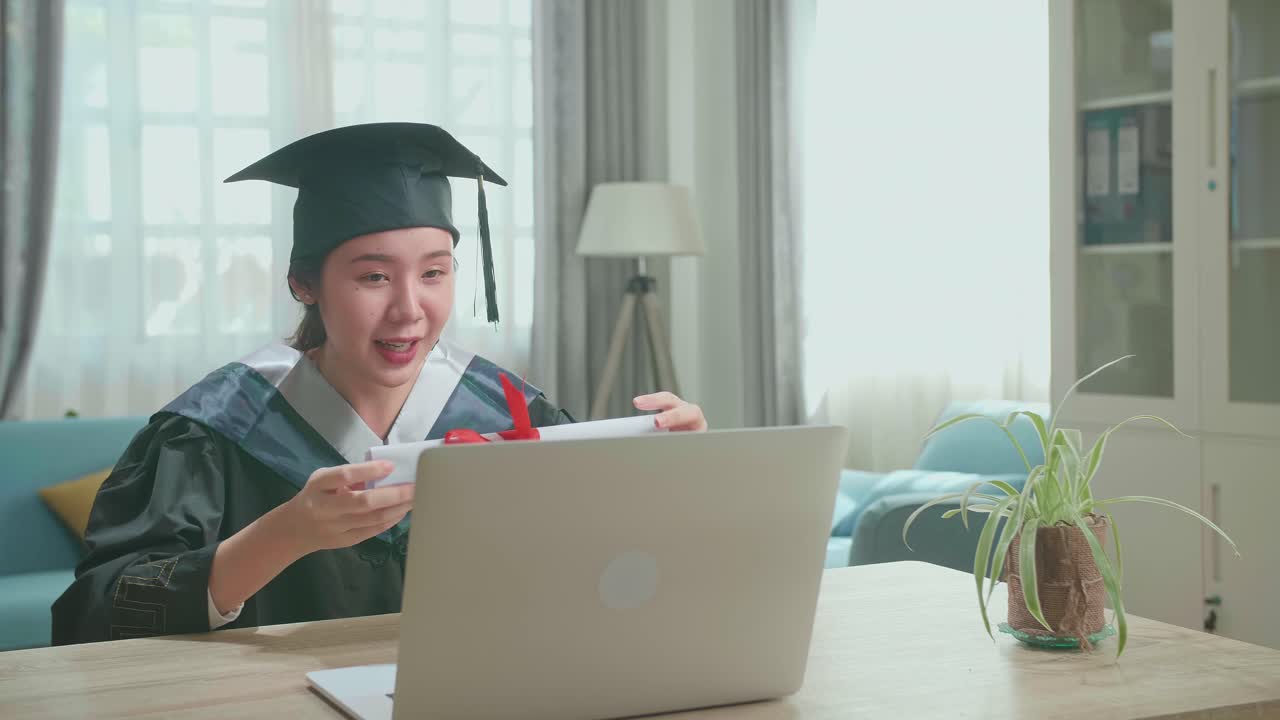 Excited Asian Woman Showing Off A University Certificate To The Family During An Online Video Call. Pretty Female Graduate Wearing A Graduation Gown And Cap Sitting On The Living Room