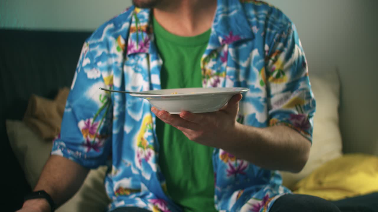 An unrecognizable young happy man seated on a sofa starts eating from a bowl full of soy milk with a spoon while watching television at night medium shot