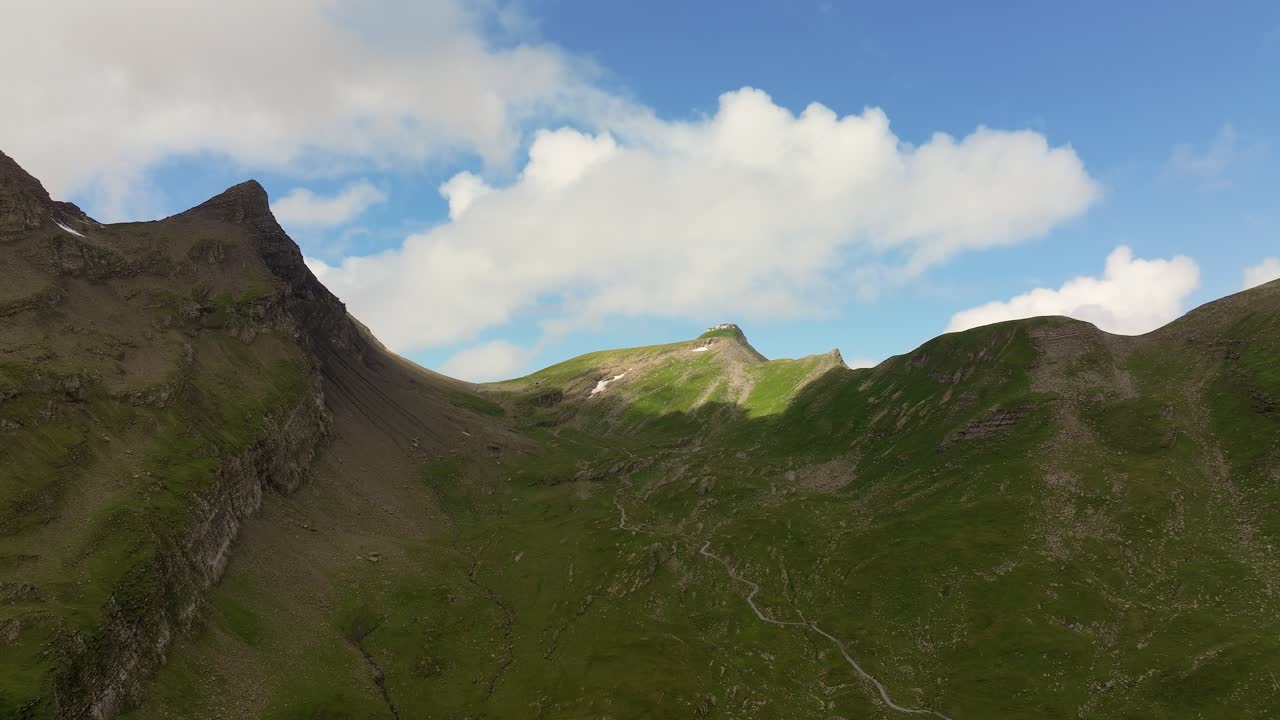 Veduta aerea di montagne alpine con ripidi pendii rocciosi e creste verdi sotto un cielo azzurro brillante, uno stretto sentiero escursionistico si snoda attraverso il terreno accidentato.